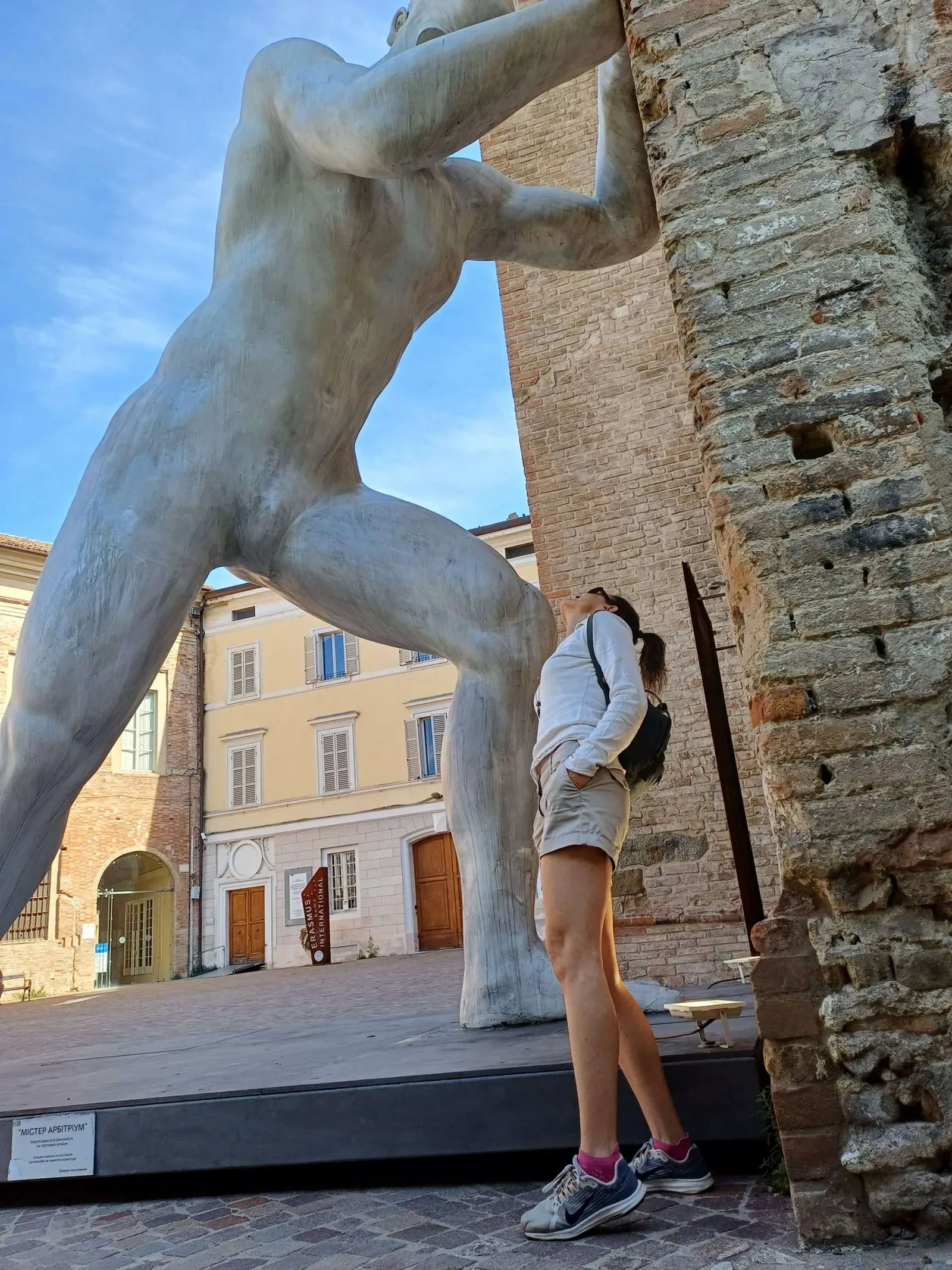 La scultura monumentale Mr. Arbitrium, un gigante intento a spingere il muro del Santuario di San Francesco del Prato, osservata dal basso da una visitatrice